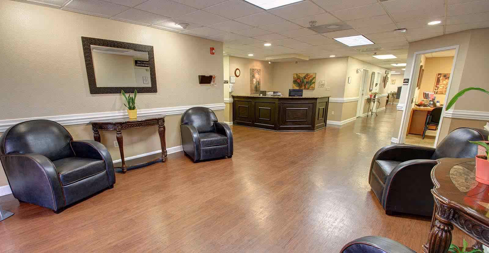 A bright assisted living lobby with black leather chairs, a reception desk, side tables with plants, and a hallway.