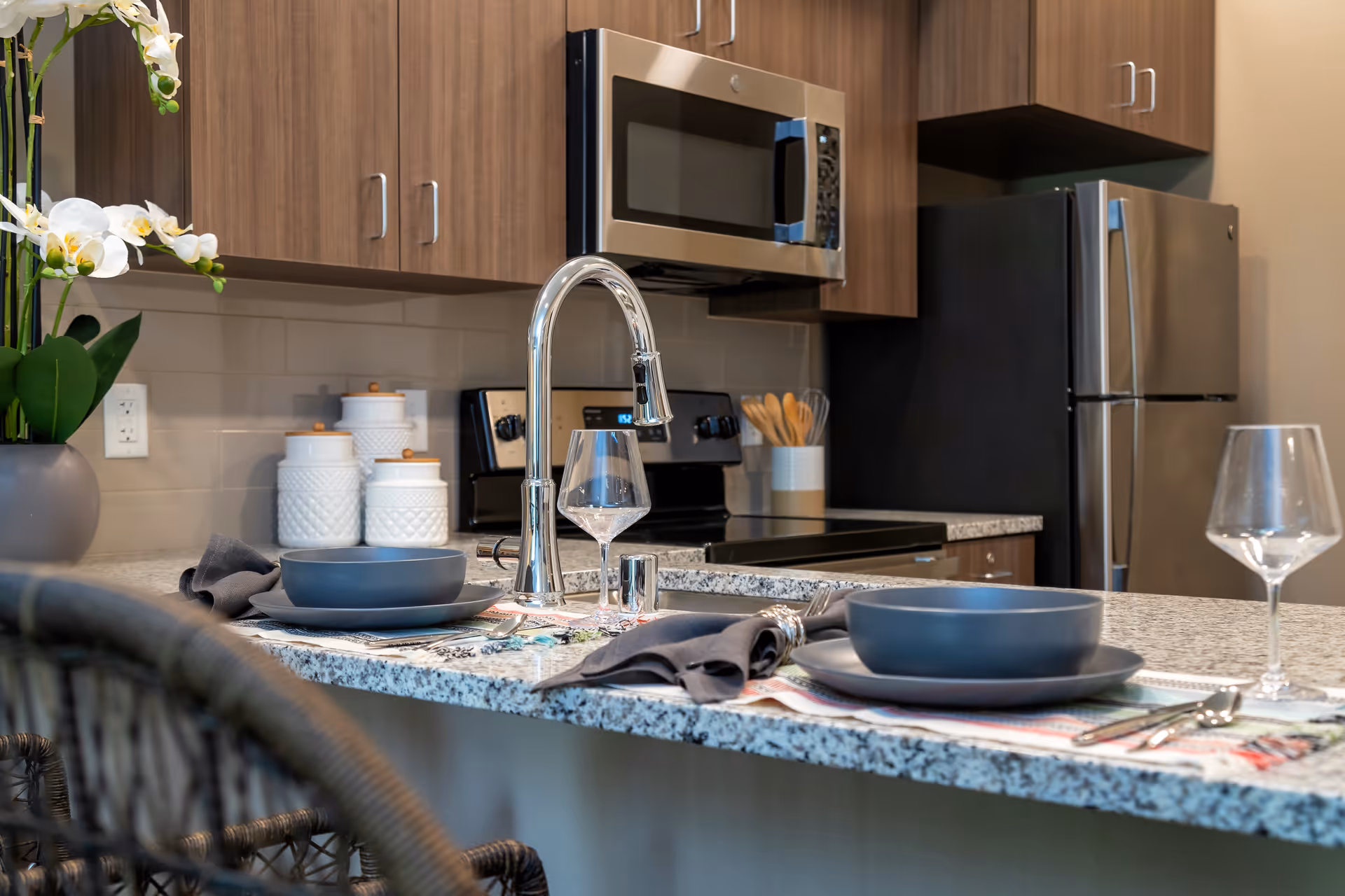 Modern kitchen with granite countertop island set for two with gray plates, bowls, napkins, and wine glasses. Stainless steel appliances including microwave, stove, and refrigerator. Wooden cabinets and a vase with white orchids on the counter.