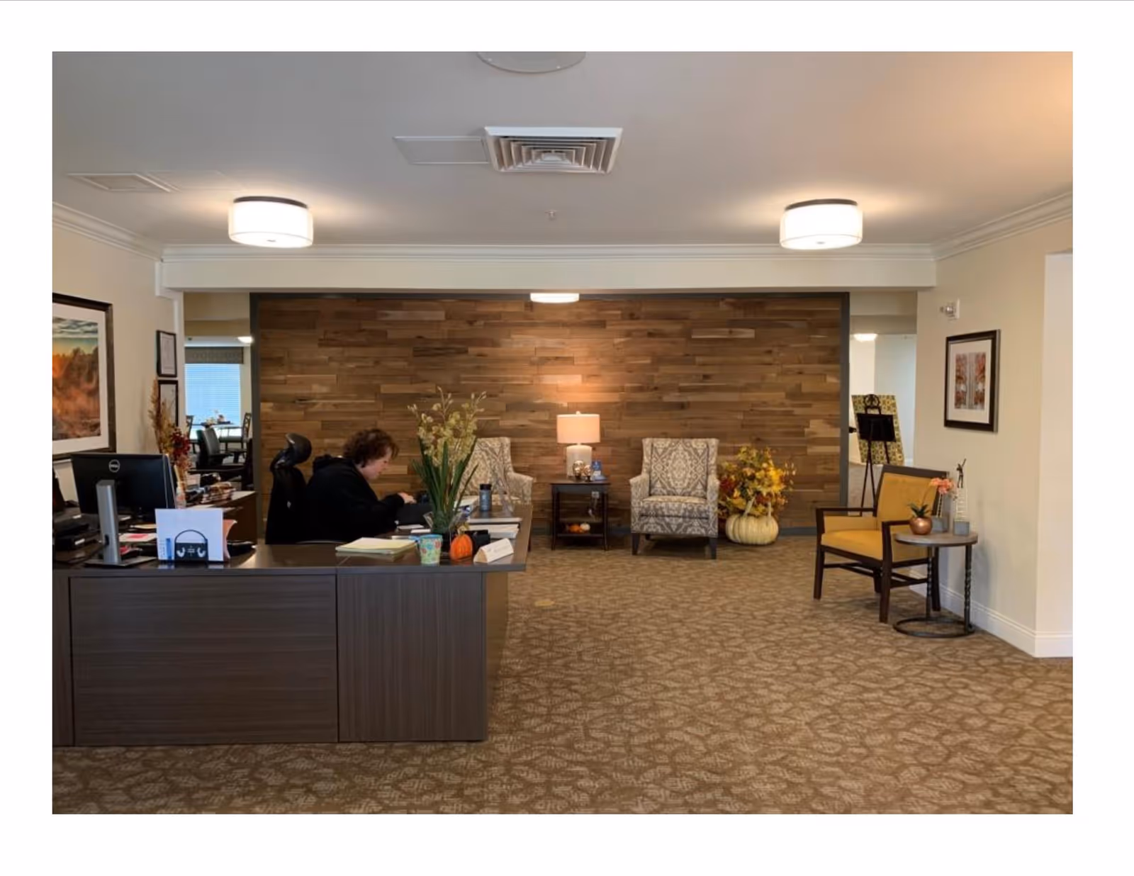 Reception area of a senior living facility with a woman seated at a dark wood desk working on paperwork. Behind the desk is a seating area with two patterned armchairs, a small table with a lamp, and decorative plants including a pumpkin. The wall behind the seating area has a wooden panel design. The carpet is patterned and the room is softly lit with ceiling lights.