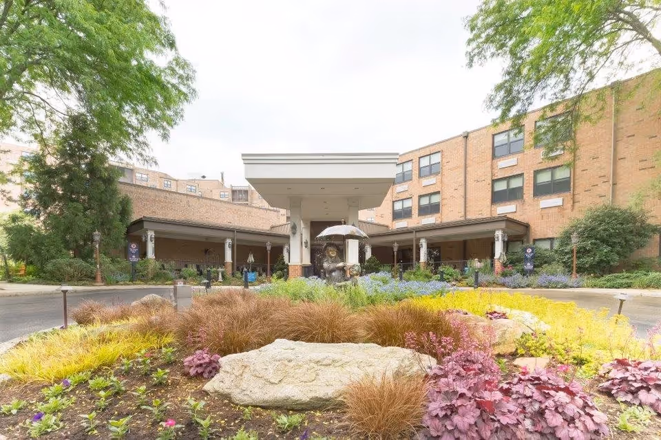 Front exterior view of The Lafayette facility showing a circular driveway with landscaped flower beds and a statue holding an umbrella in the center. The building is a multi-story brick structure with large windows and a covered entrance.