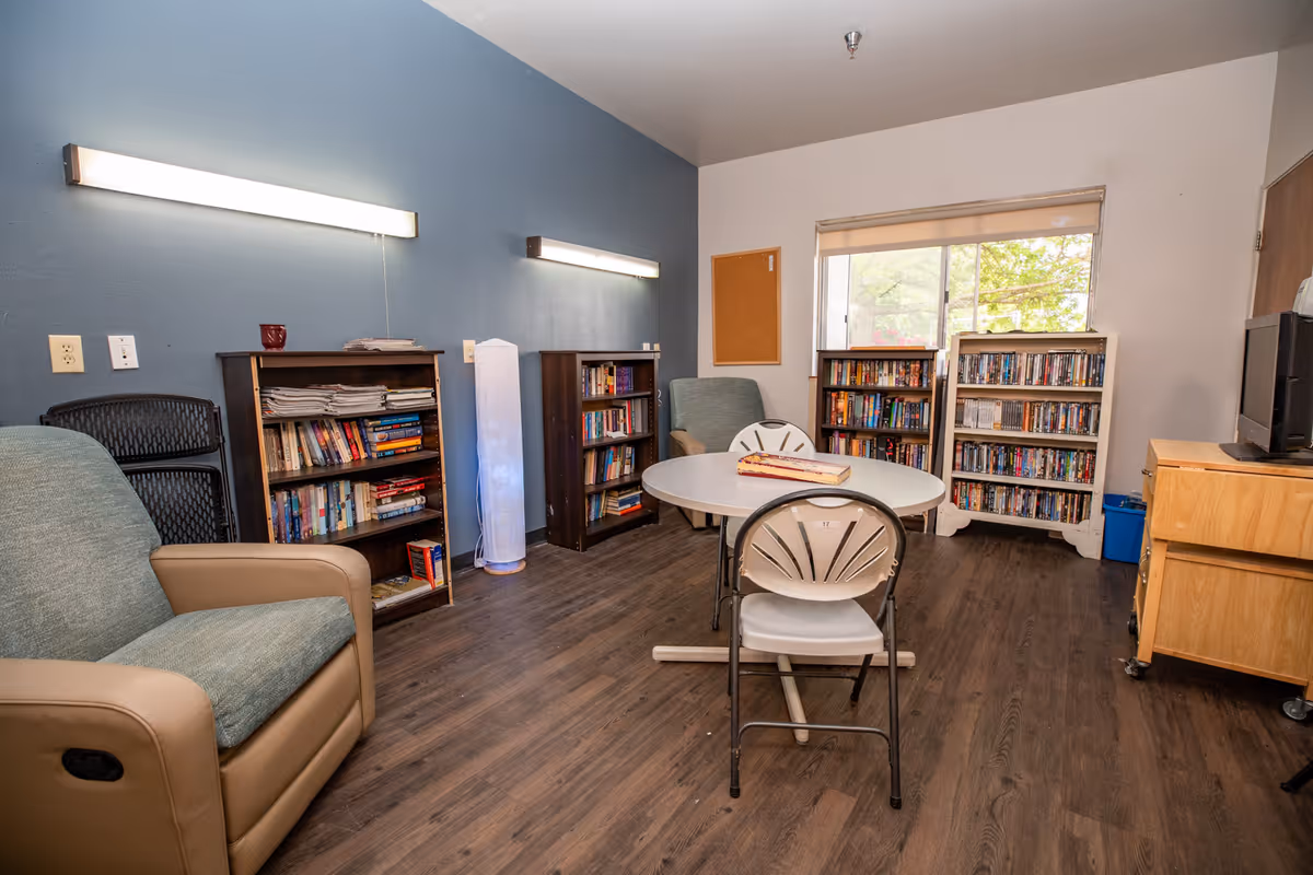 A cozy interior room with a round table and four chairs in the center. There are bookshelves filled with books and magazines against the walls, a comfortable armchair, a tall floor lamp, and a window letting in natural light. The walls are painted blue and white, and the floor has dark wood-style flooring.