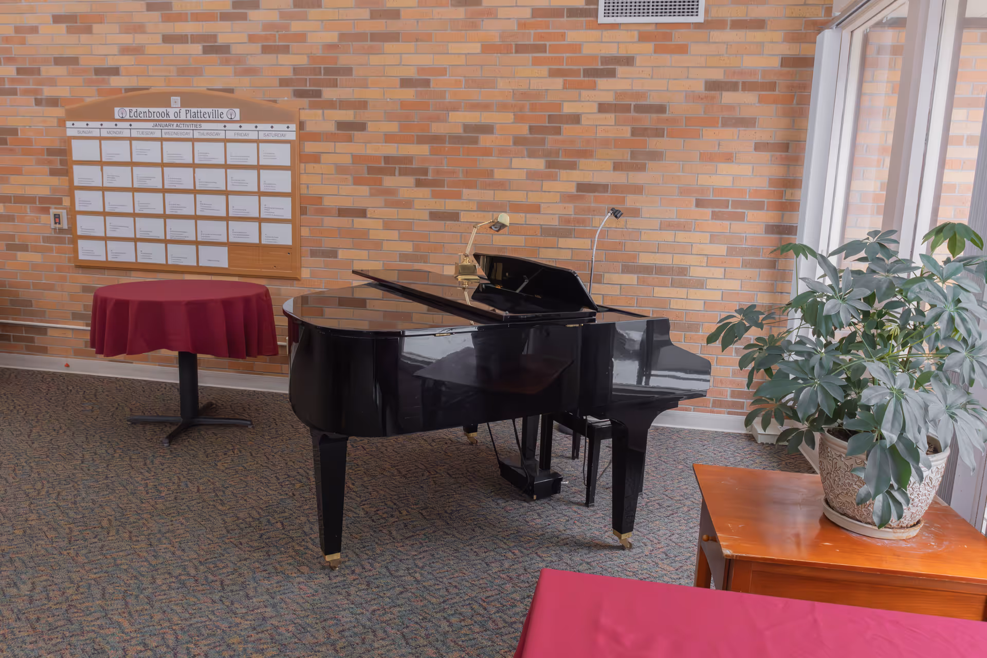 Interior room with a black grand piano, two small round tables covered with red tablecloths, a large potted plant on a wooden table, and a brick wall with a bulletin board displaying the January activities schedule for Edenbrook of Platteville.