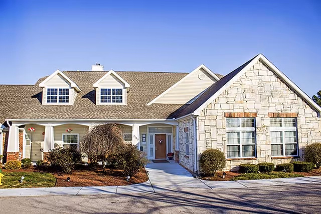 Exterior view of a single-story residential-style building with a stone and siding facade, multiple windows, a front porch with columns, and a driveway leading to the entrance under a clear blue sky.