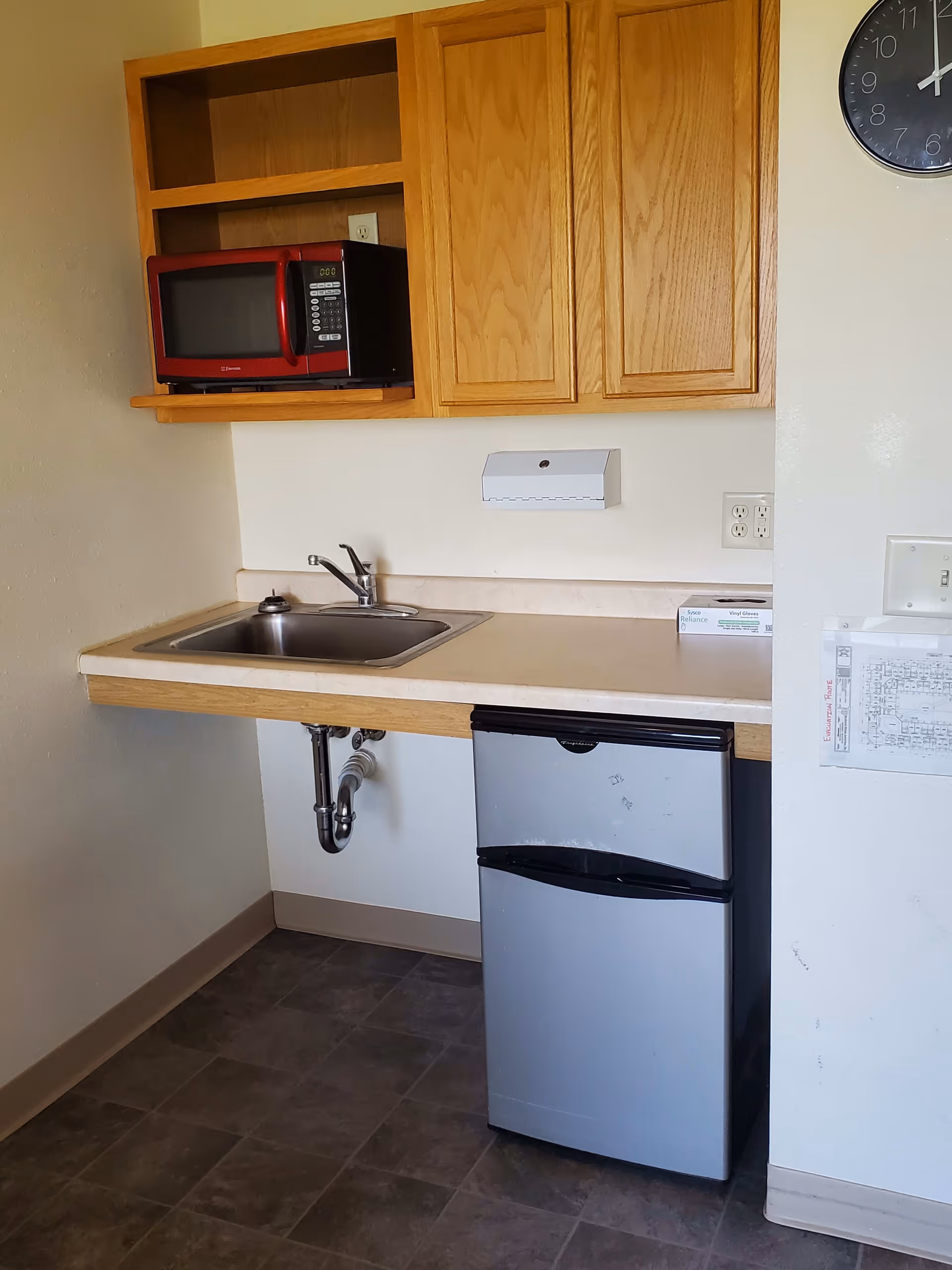 Small kitchenette area with a stainless steel sink, wooden cabinets above, a red microwave on a shelf, a small silver refrigerator below the counter, and a wall clock showing 12:05.