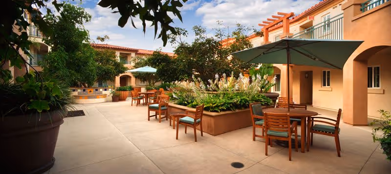 Outdoor courtyard area at White Sands La Jolla featuring wooden tables with green umbrellas, surrounded by potted plants and greenery, with a beige building in the background under a partly cloudy sky.