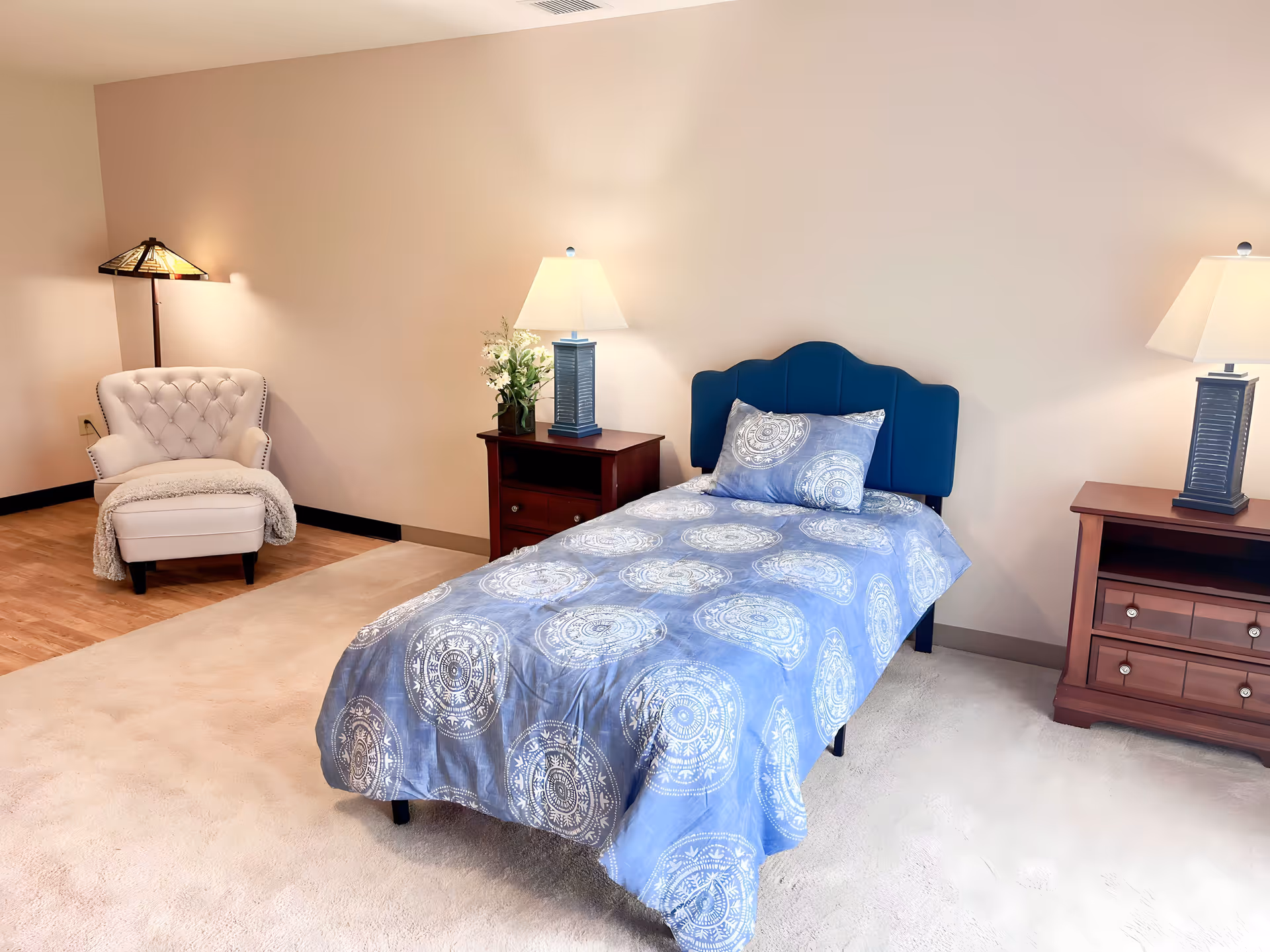 Neatly furnished bedroom with a single bed covered in blue patterned bedding, two nightstands with lamps, and an upholstered armchair in the corner.