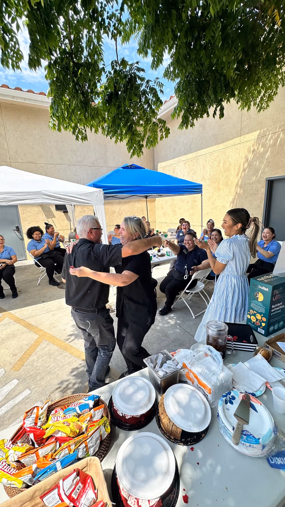 A group of people gathered outdoors in a courtyard area with beige walls and a tree providing shade. Two people are dancing in the center while others sit on folding chairs under blue and white canopies, clapping and smiling. In the foreground, there is a table with snacks, plates, napkins, and a blender box.