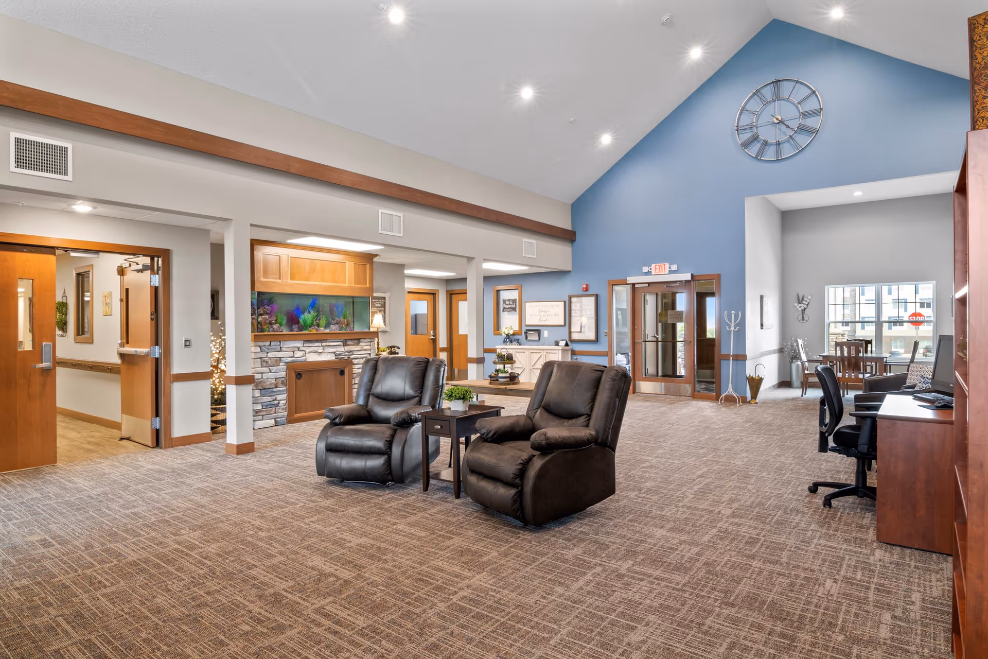Spacious senior living facility common area with two black recliner chairs and a small table in the center. The room features a high vaulted ceiling painted blue with a large wall clock, a stone fireplace with an aquarium above it, and multiple wooden doors leading to other rooms. There is a desk with a computer and office chair on the right side, and a seating area with a table and chairs near large windows in the background.