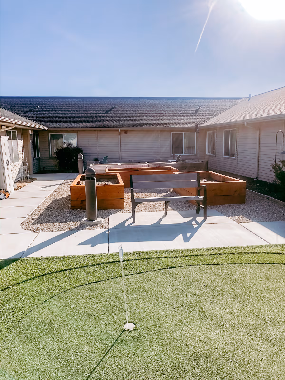 Outdoor courtyard area at Copper Springs Senior Living featuring a small putting green with a hole and flag, a bench, raised wooden garden beds, and a building with multiple windows surrounding the courtyard under a clear blue sky.