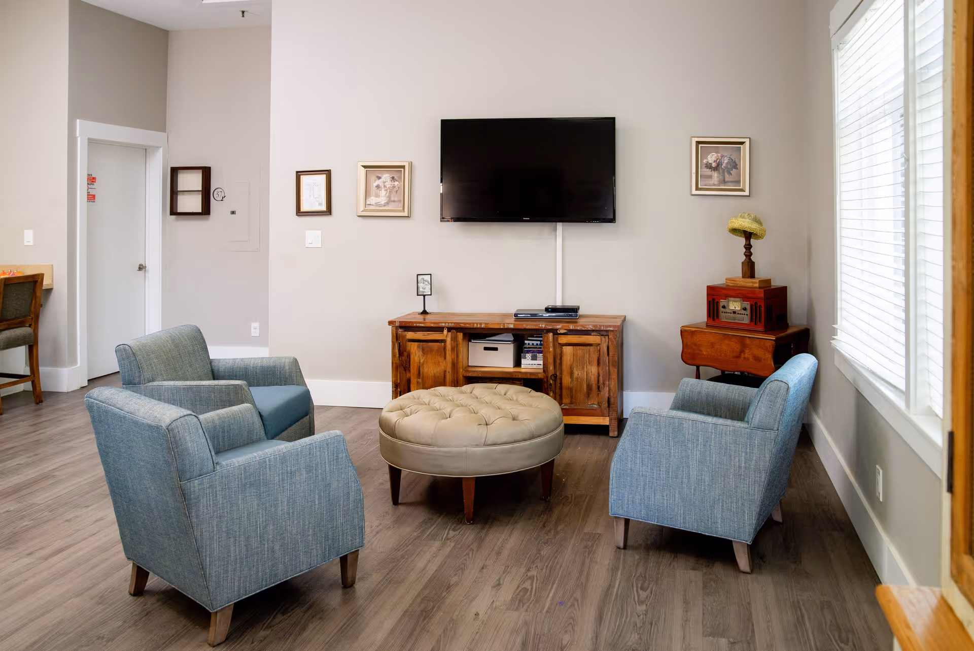 A cozy living room area with three blue upholstered armchairs arranged around a round beige tufted ottoman. A wooden TV stand with storage compartments holds a flat-screen TV mounted on the wall above it. The walls are decorated with framed artwork, and a window with white blinds allows natural light to enter the room. The flooring is wood, and there is a small wooden side table with a decorative lamp on it.