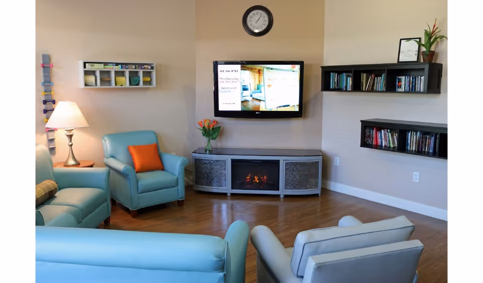 A cozy living room with light blue armchairs and a sofa arranged around a modern electric fireplace. A flat-screen TV is mounted on the wall above the fireplace, displaying a schedule. There are two wall-mounted bookshelves filled with books, a table lamp on a side table, a clock above the TV, and a vase with flowers on the fireplace stand. The room has wooden flooring and beige walls.