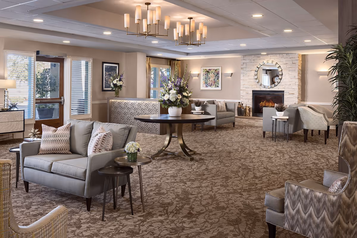 A spacious and elegantly decorated living room area in a senior living facility featuring multiple seating arrangements with sofas and armchairs, a round table with a floral centerpiece, a stone fireplace with a circular mirror above it, framed artwork on the walls, large windows with curtains, and soft lighting from ceiling fixtures and wall sconces.