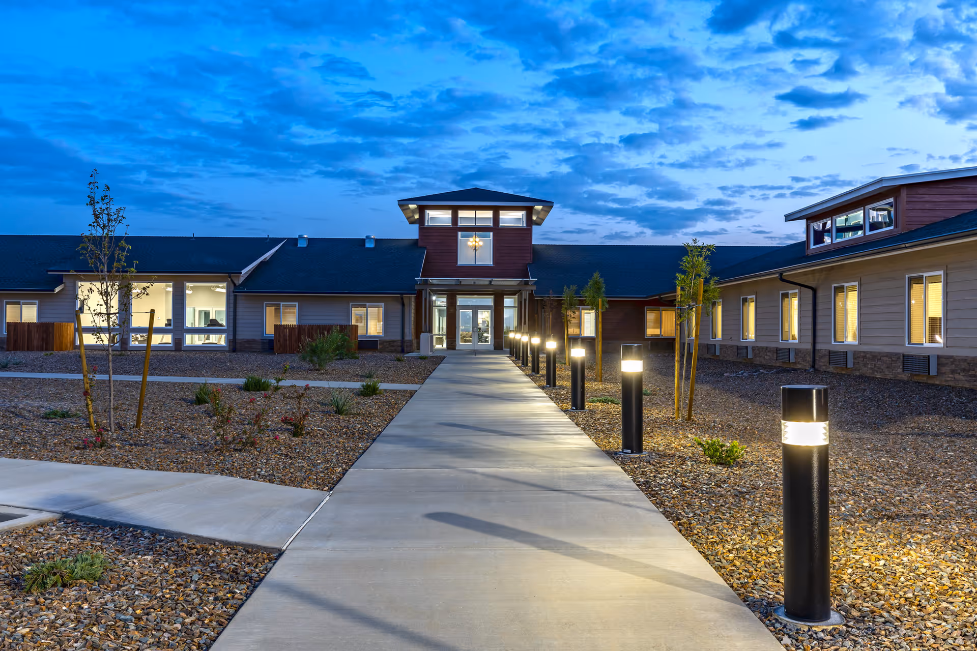 Evening view of the entrance walkway to The Landings Of Prescott Valley facility, with illuminated pathway lights, landscaped gravel areas with small plants and trees, and a building with multiple windows and a central tower under a partly cloudy sky.