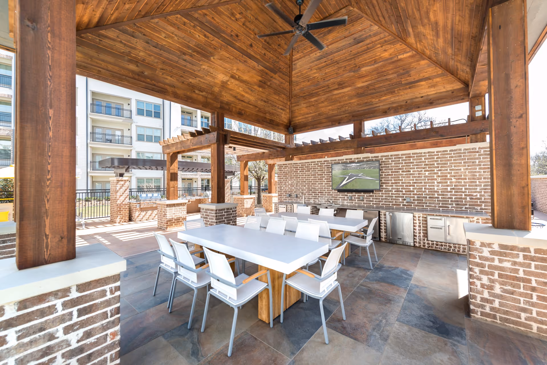 Outdoor covered patio area with wooden ceiling and brick pillars, featuring two white tables with white chairs, a ceiling fan, a mounted TV on a brick wall, and an outdoor kitchen with stainless steel appliances.