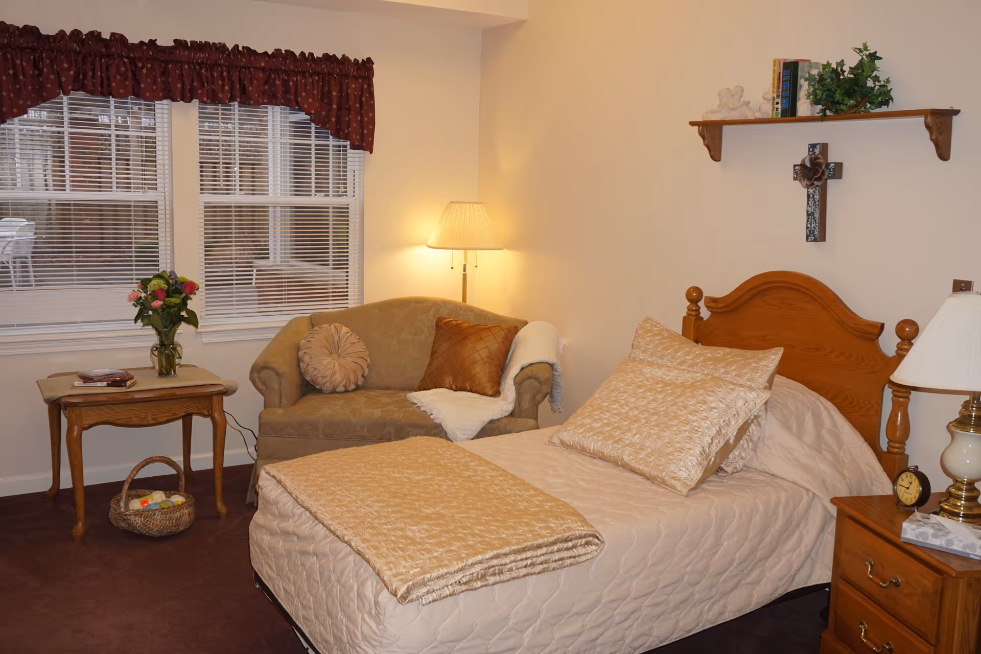 A cozy bedroom in a senior living facility featuring a single wooden bed with beige quilted bedding and pillows. Next to the bed is a wooden nightstand with a lamp, an alarm clock, and a tissue box. A small beige loveseat with decorative pillows and a throw blanket is positioned near a window with white blinds and a maroon valance. A wooden side table holds a vase of flowers and some books, with a basket underneath. A wall shelf above the bed displays books, a plant, and decorative items, including a cross.