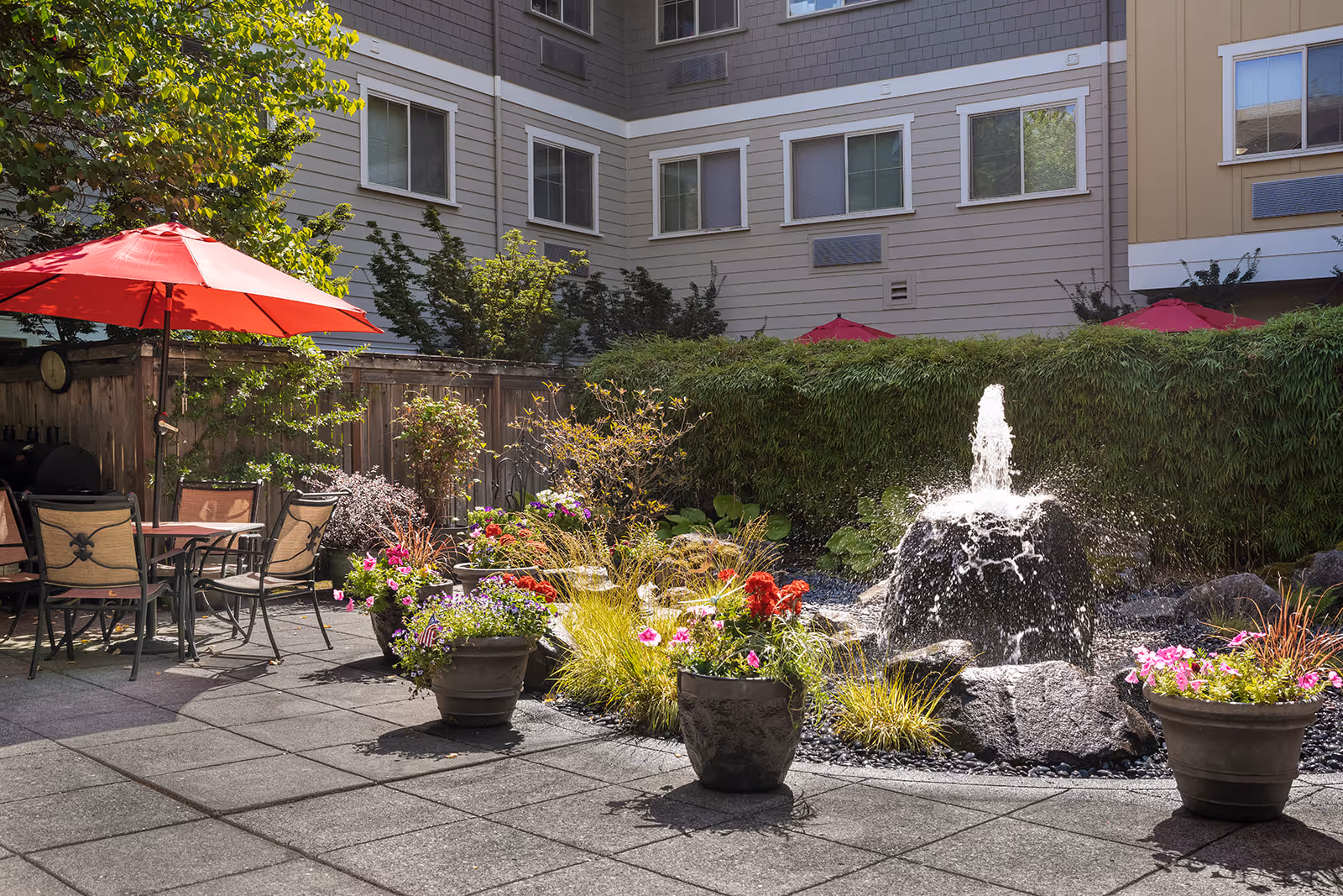 Outdoor patio area with a stone water fountain surrounded by plants and flowers in pots. There are tables and chairs with red umbrellas providing shade. The background shows the exterior walls and windows of a multi-story building.