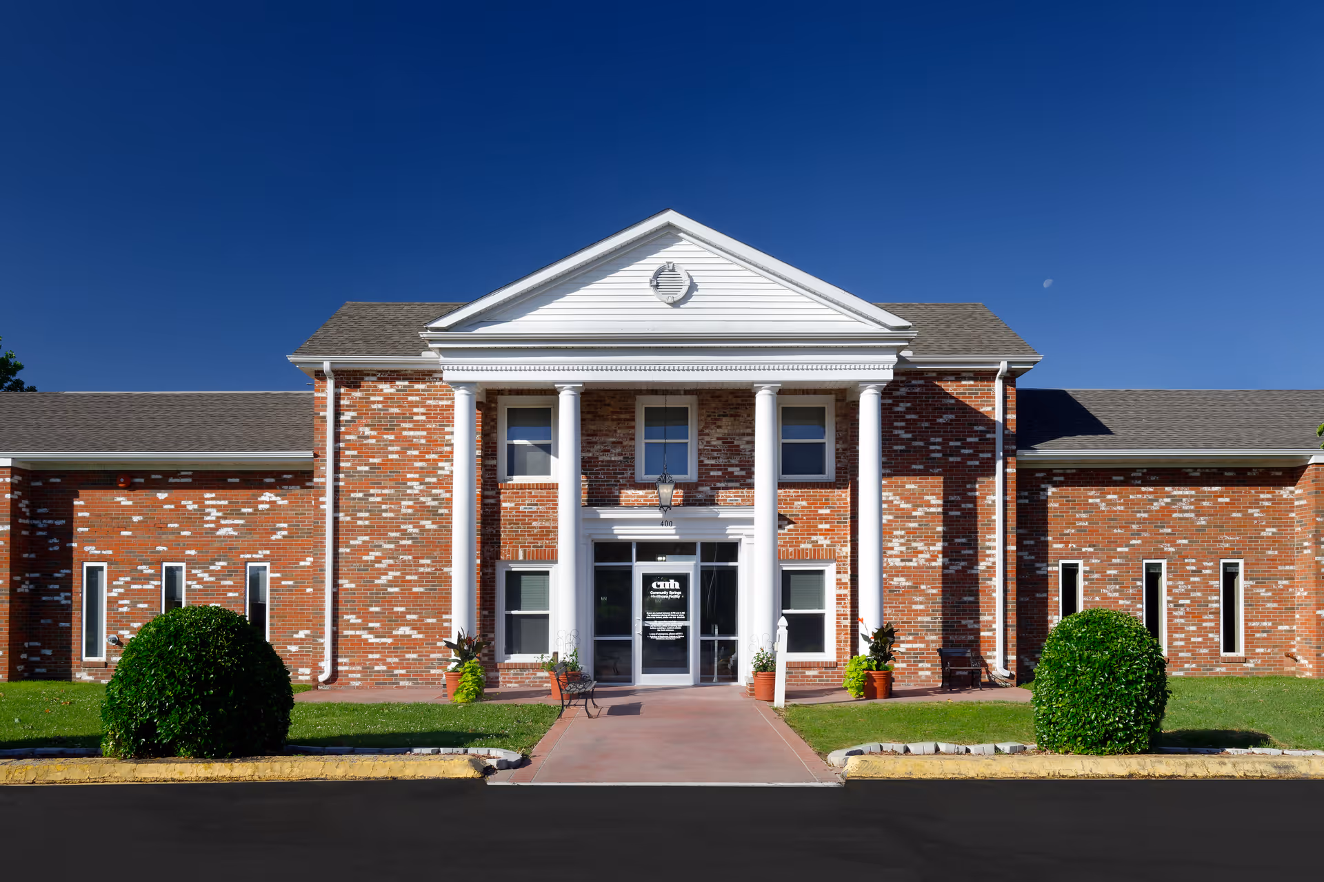 Front exterior view of a brick healthcare facility building with white columns and a triangular pediment above the entrance. There are two bushes on either side of the walkway leading to the glass double doors. The sky is clear and blue.