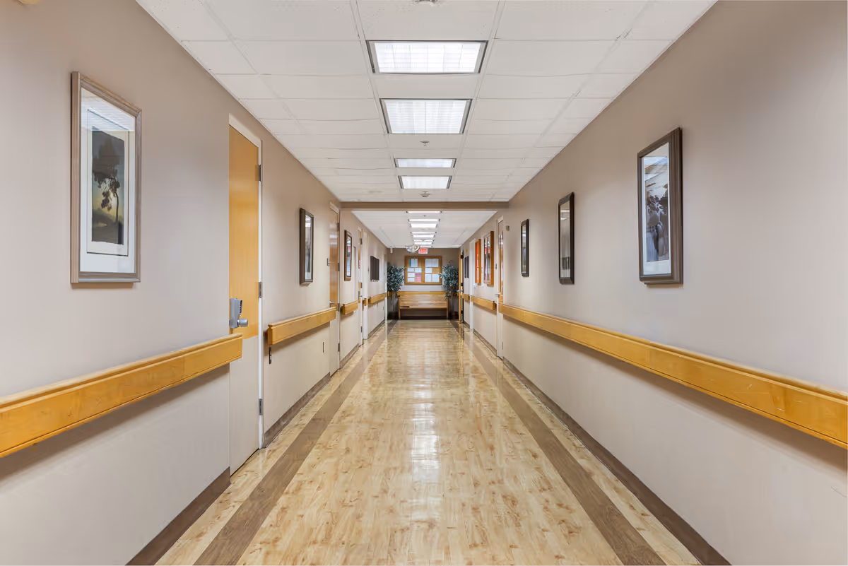 A long, clean hallway in a senior living facility with light wood flooring, beige walls, handrails on both sides, several closed doors, framed pictures on the walls, and fluorescent ceiling lights.