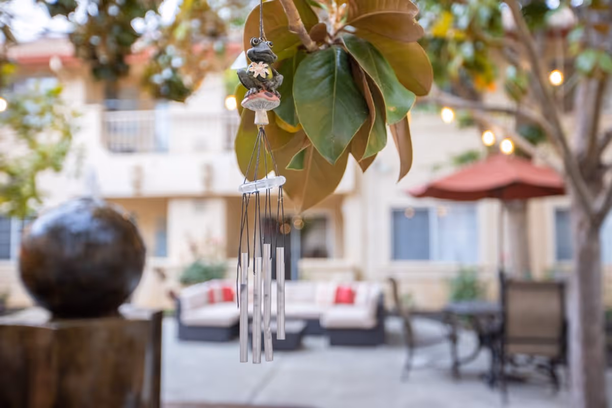 Close-up of a decorative wind chime hanging from a tree branch with large green leaves. In the blurred background, there is an outdoor seating area with cushioned chairs, a table with an umbrella, and a building facade with windows.