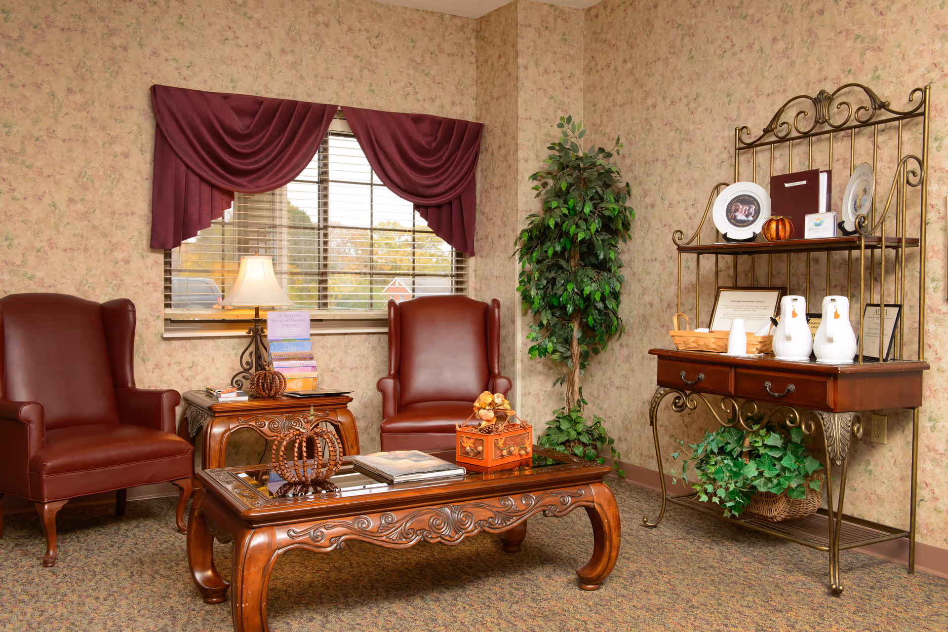 A cozy sitting area with two brown leather armchairs, a wooden coffee table with decorative items, a side table with a lamp and books, a window with maroon curtains, a tall green plant, and a wooden and metal shelving unit holding white pitchers, framed certificates, and decorative plates.