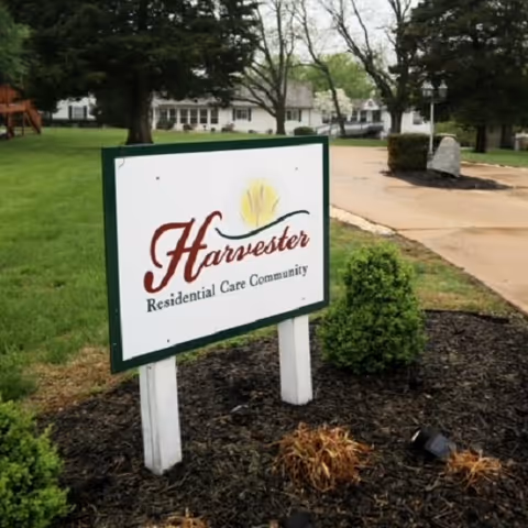 A sign for Harvester Residential Care Community is displayed on a grassy area near a paved driveway. The background shows trees and a white building, likely part of the care facility.