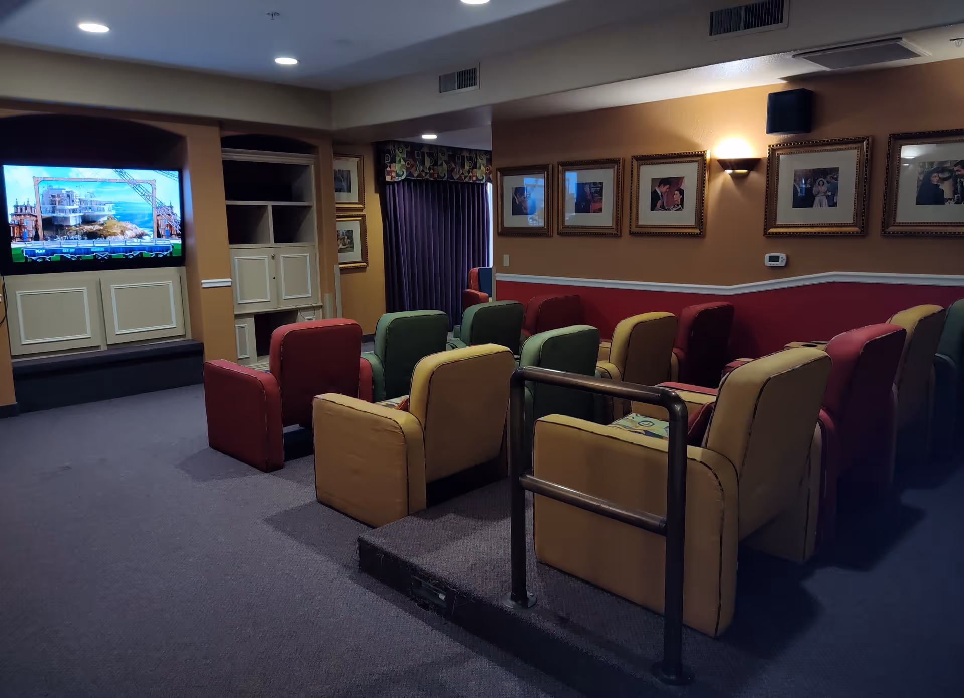 A small theater room with rows of colorful cushioned chairs facing a large flat-screen TV mounted on the wall. The room has framed pictures on the walls, a purple curtain, and a handrail in front of the seating area.
