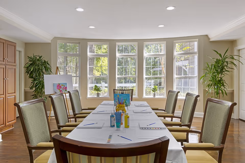 A bright and spacious activity room with a long table covered in a white tablecloth, set up for painting with brushes, paints, and paper. The room has large windows letting in natural light, green plants on the windowsills and in the corners, and wooden chairs with green cushions arranged around the table.