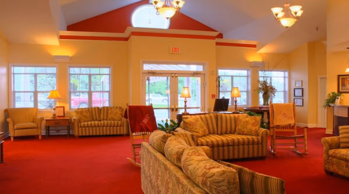 A spacious and warmly lit living room area with red carpet, multiple striped and floral patterned sofas, armchairs, rocking chairs with red blankets, several table lamps, large windows, and a glass door entrance under a high ceiling with a chandelier.
