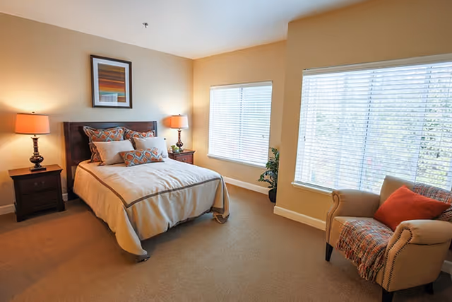 A cozy bedroom with a neatly made bed featuring beige and orange patterned pillows and a beige comforter. There are two bedside tables with lamps on either side of the bed, a framed abstract artwork above the headboard, two large windows with white blinds, a potted plant in the corner, and a beige armchair with an orange pillow and a throw blanket near the windows.