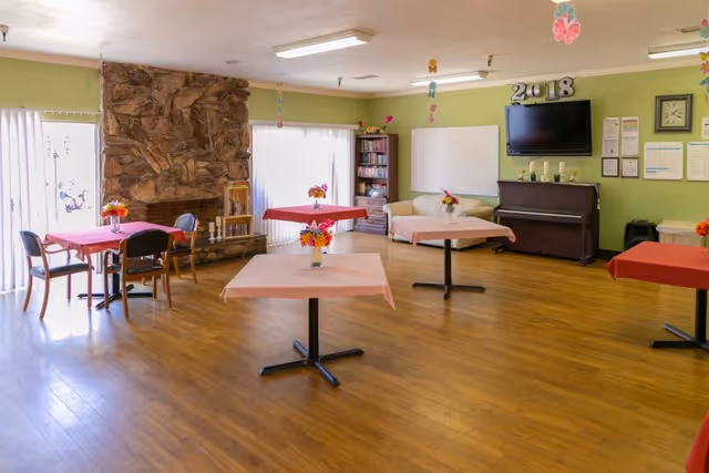A spacious room with wooden flooring and light green walls featuring several tables covered with pink and red tablecloths, each decorated with a vase of colorful flowers. There is a stone fireplace on the left side, a bookshelf with books, a piano with candles and a TV mounted above it, and a whiteboard on the wall. The room is well-lit with natural light coming through large windows with vertical blinds, and ceiling lights. Decorative butterflies hang from the ceiling.