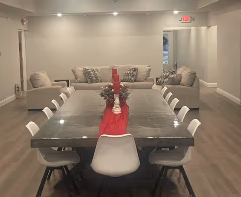 Communal dining room with a long table surrounded by white chairs, a red runner and candles down the center, and sofas in a lounge area at the back.