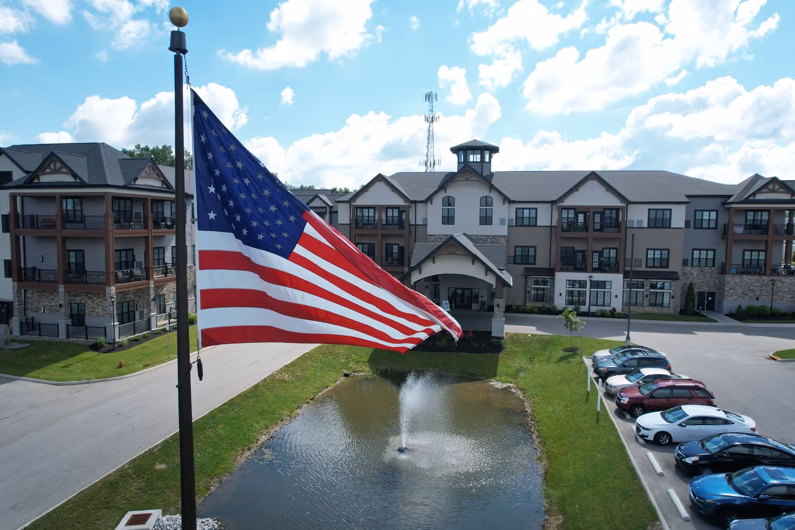 Exterior view of a senior living facility with a large American flag in the foreground, a pond with a fountain, a parking lot with cars, and a multi-story building with balconies under a partly cloudy sky.