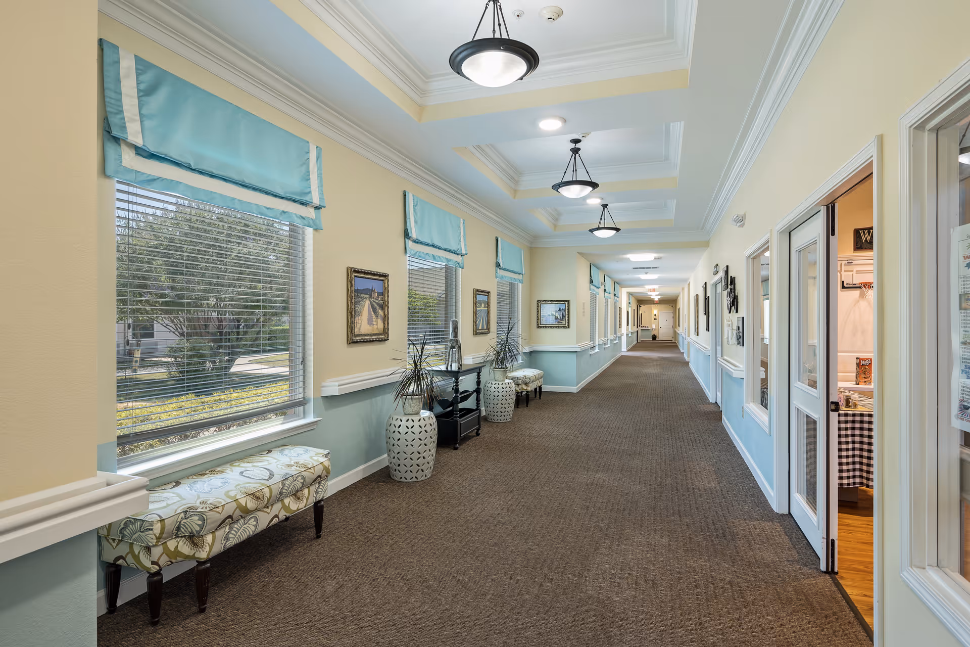 Long carpeted interior hallway with windows, benches, decorative plants, and ceiling lights in a senior living facility.