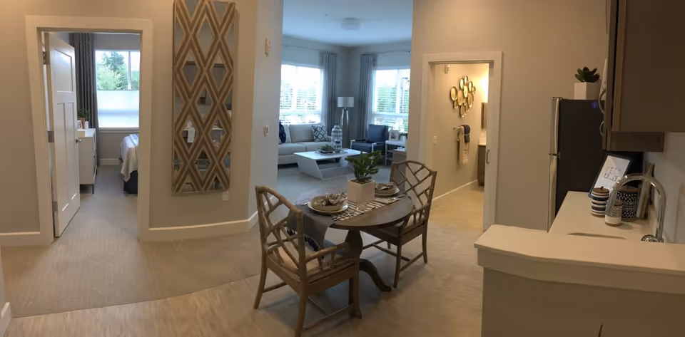 Open-plan interior of an assisted living apartment with a small dining table in the foreground, kitchen counter to the right, and a living room and bedroom visible in the background.