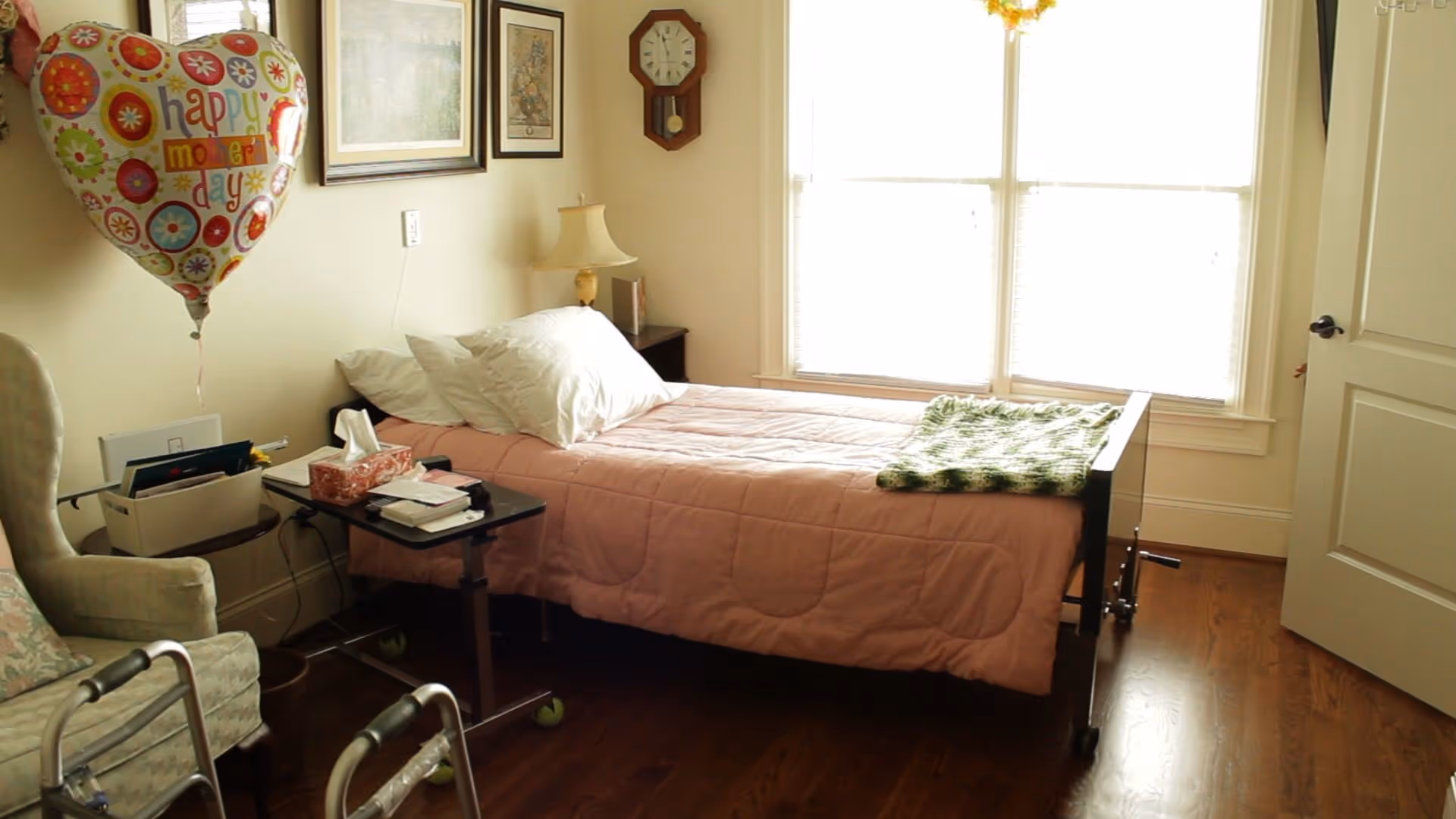 A small bedroom with a single bed covered in a pink quilt and white pillows. A green knitted blanket is folded at the foot of the bed. Next to the bed is a small black overbed table with tissues and other items. A beige armchair with a floral cushion is partially visible on the left. A walker is in front of the chair. On the wall above the bed are two framed pictures and a wooden clock. A heart-shaped balloon with 'happy mother's day' written on it is floating near the wall. The room has wooden flooring and a large window with white blinds letting in bright light. A closed white door is on the right side.