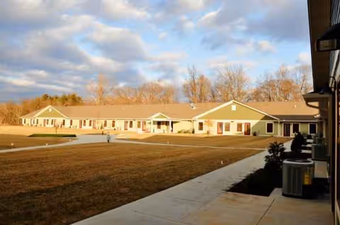 Single-story senior living building front with walkway, lawn, and leafless trees in late afternoon light.