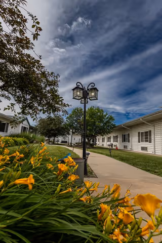 Outdoor walkway at Bailey Pointe at Plattsmouth with yellow flowers in the foreground, a black lamp post in the center, and white residential buildings on either side under a partly cloudy sky.