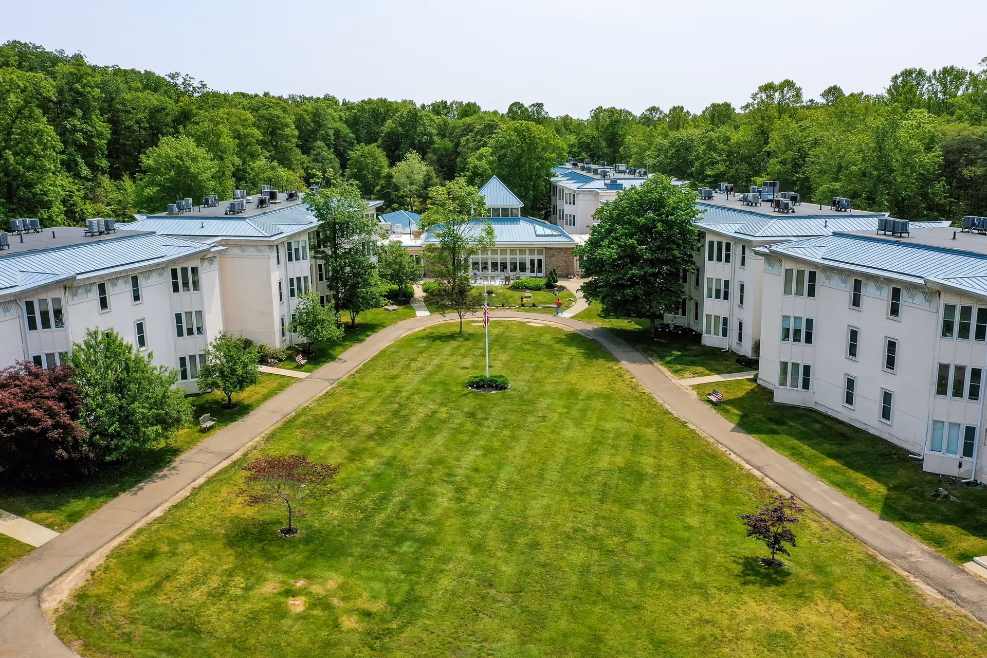 Aerial view of Guilford Retirement Residence showing multiple white buildings with blue roofs surrounding a large green lawn with a flagpole in the center. The area is bordered by trees and has paved walkways and benches along the sides.