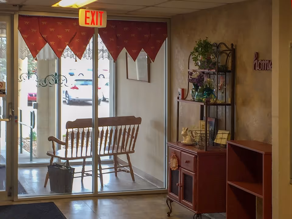 Interior view of an entrance area with glass doors leading outside. A wooden bench is placed near the doors, and a small basket is on the floor next to it. To the right, there is a red cabinet with decorative items and plants on top and a metal shelf above it. An exit sign is visible above the doors.