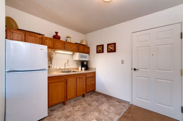 Small kitchenette with wooden cabinets, a white refrigerator, sink, microwave, and a door on the right.