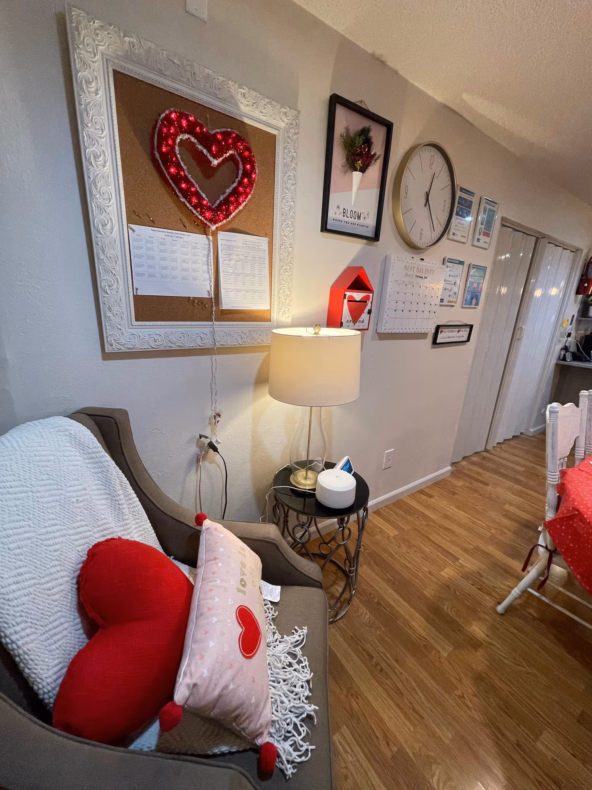 Cozy living area with an armchair topped with heart-shaped pillows, a side table and lamp, and a wall decorated with a clock, calendar and framed art.
