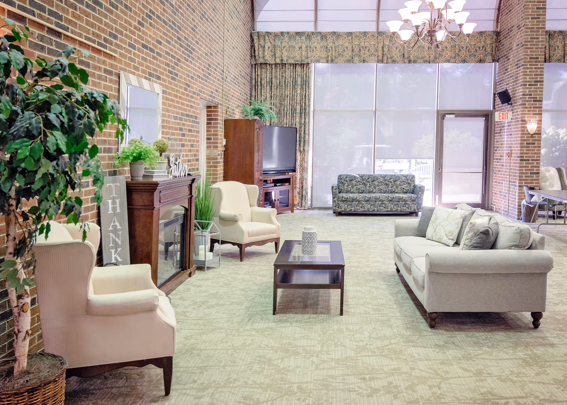 A spacious and well-lit living room area with brick walls, large windows covered by curtains, and a glass door. The room features a beige sofa with cushions, two beige armchairs, a dark wooden coffee table with a decorative candle, a TV on a wooden stand, a fireplace with plants and decor on the mantel, and a large potted plant on the left side.