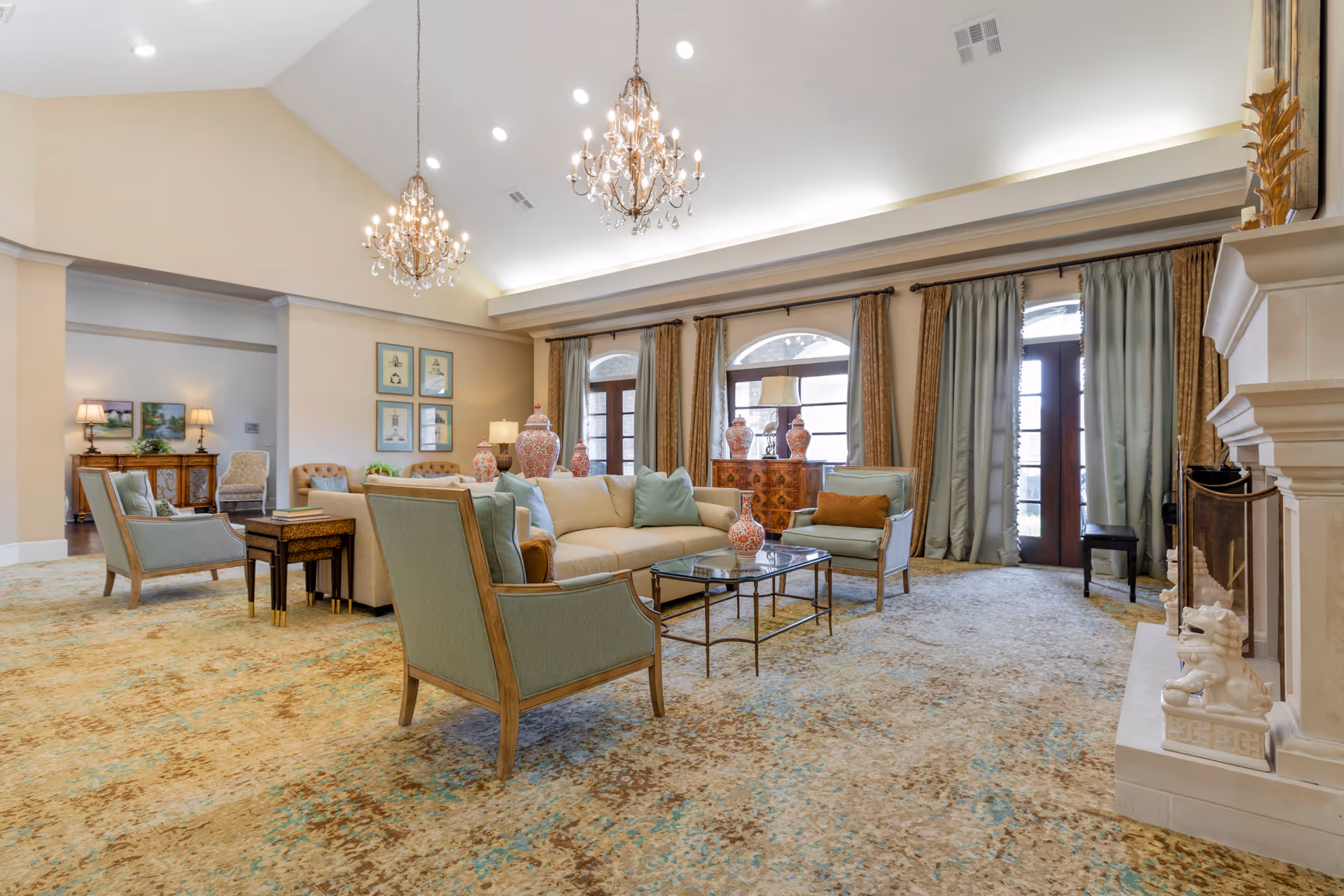 A spacious and elegantly decorated living room in a senior living facility featuring a beige sofa with light blue and brown cushions, two light blue armchairs, a glass coffee table with decorative vases, large windows with curtains, two chandeliers hanging from a high ceiling, and a fireplace with decorative sculptures.