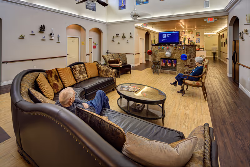A cozy assisted living facility common area with two elderly residents seated on comfortable sofas and chairs facing a stone fireplace with a mounted TV above it. The room features wood flooring, decorative pillows, wall art, and a glass coffee table with magazines.