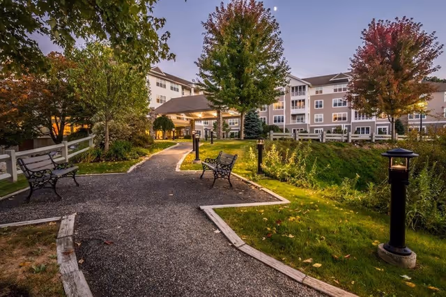 Outdoor pathway in a senior living facility with benches, trees with autumn foliage, lamp posts, and a multi-story building in the background under a twilight sky.