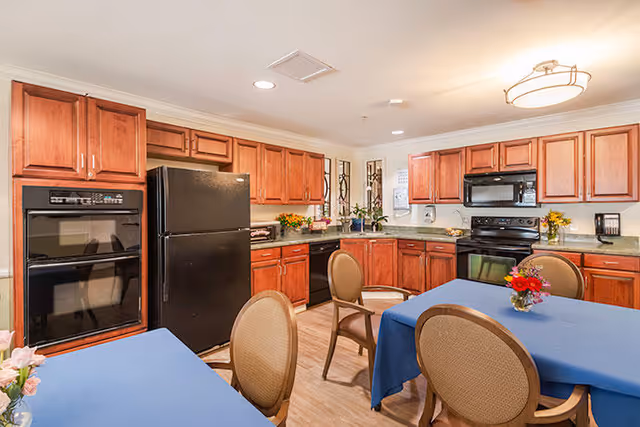 A well-lit kitchen with wooden cabinets, a black refrigerator, oven, and microwave. There are two tables covered with blue tablecloths, each surrounded by chairs. The kitchen has a light wood floor and a ceiling light fixture. Flowers in vases are placed on the tables and countertops.