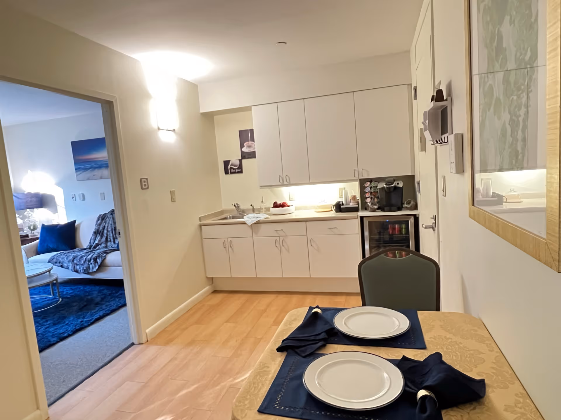 A small dining area with a table set for two with white plates and dark blue napkins. Behind the table is a kitchenette with white cabinets, a sink, a coffee maker, and a mini fridge. To the left, an open doorway leads to a living room with a white couch, blue pillows, a blue rug, and a side table with a lamp.