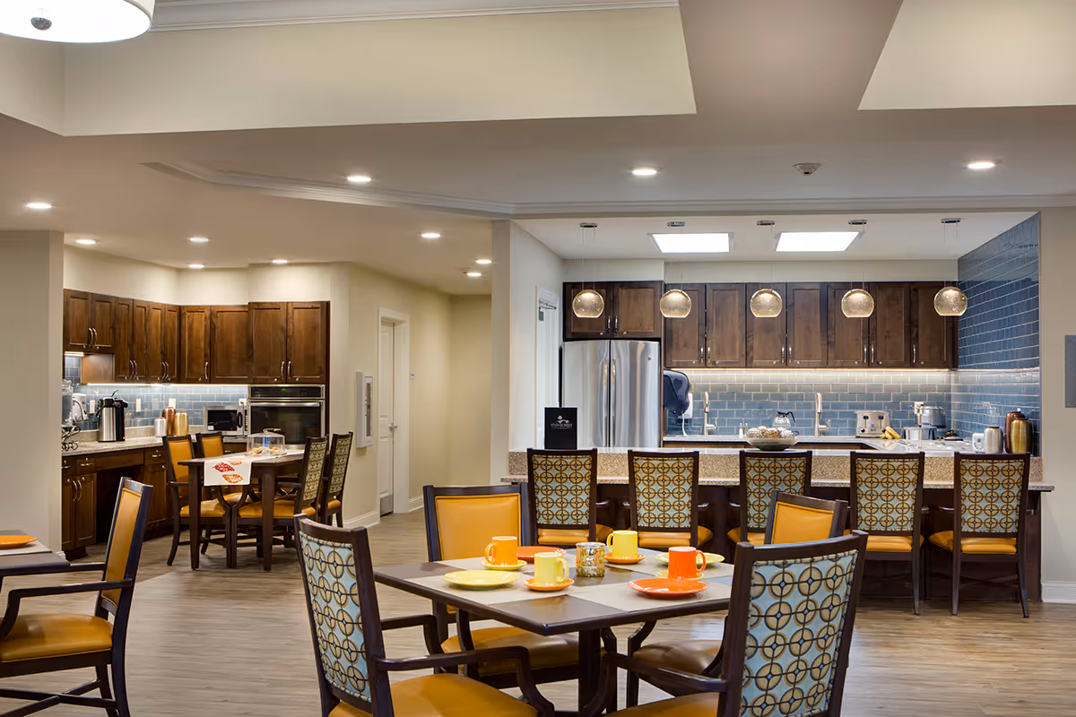 A spacious and well-lit kitchen and dining area in a senior living facility. The kitchen features dark wooden cabinets, a stainless steel refrigerator, a microwave, and a coffee maker. There is a long counter with six high chairs, and in the foreground, there are two dining tables with patterned chairs and colorful plates and mugs set on them. The floor is wooden, and the walls have a light beige color with blue tiled backsplash in the kitchen area. Pendant lights hang above the kitchen counter.