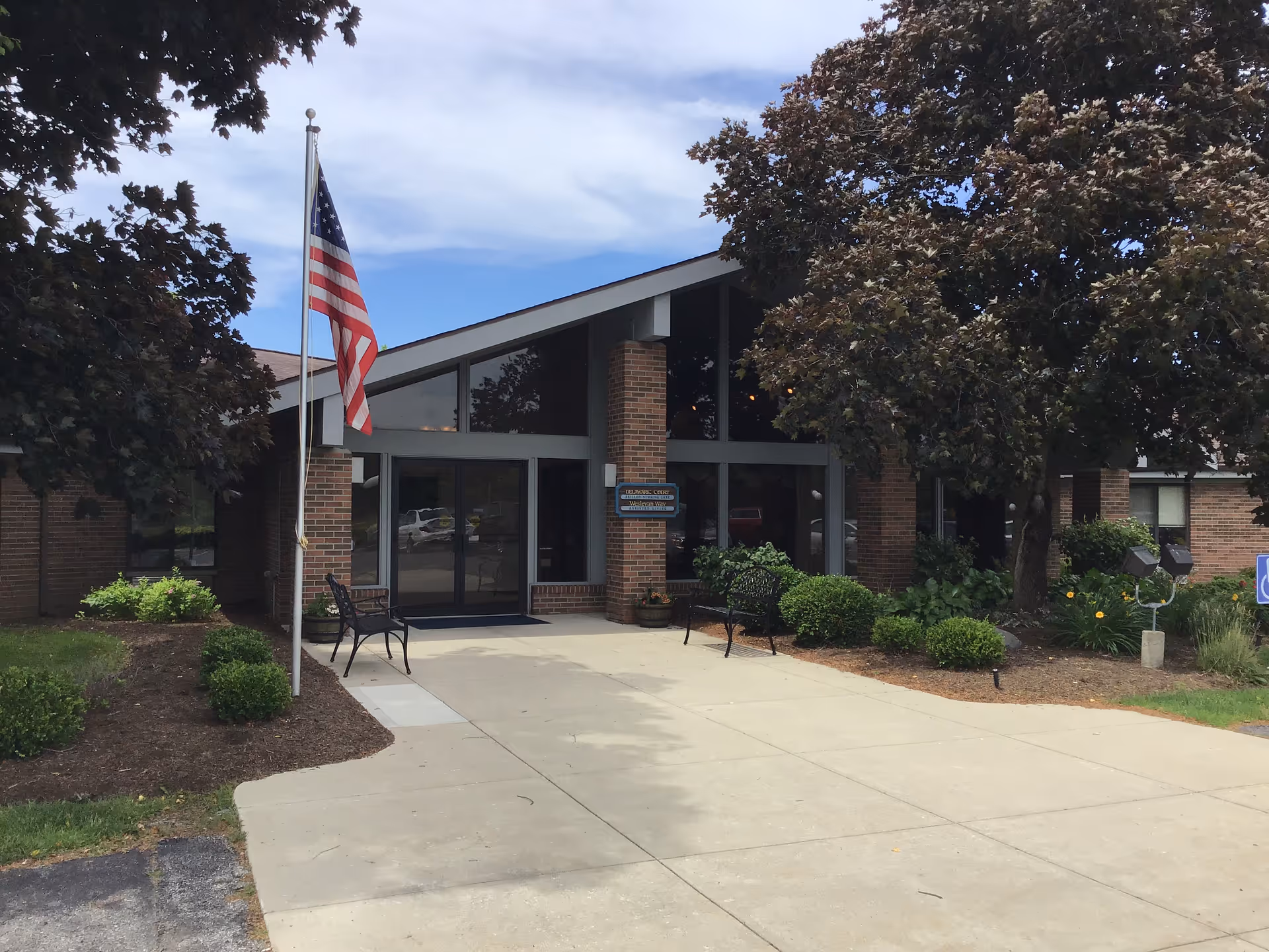 Front exterior view of Delaware Court Healthcare Center showing a brick building with large glass windows, an American flag on a flagpole, benches, and landscaped bushes and trees under a partly cloudy sky.