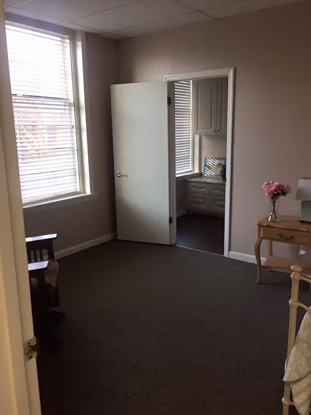 Interior view of a room with beige walls and carpeted floor. A window with white blinds is on the left wall, and a white door is open, leading to another room with cabinets and a window. A wooden side table with a vase of pink flowers and a lamp is on the right side.